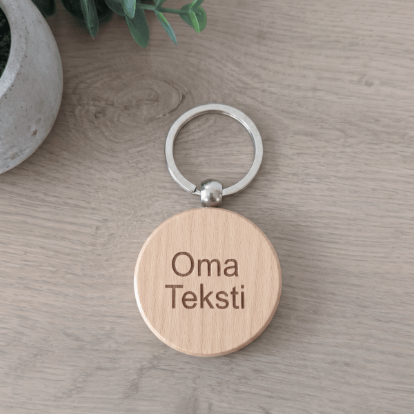 Vaaleasta puusta valmistetussa pyöreässä avaimenperässä näkyy kaiverrettu teksti Oma Teksti ja metallinen rengas luonnonsävyisellä taustalla.